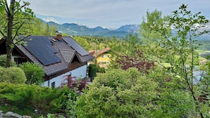 Une maison avec un toit en pente, entourée d’une végétation luxuriante et avec des montagnes en arrière-plan.