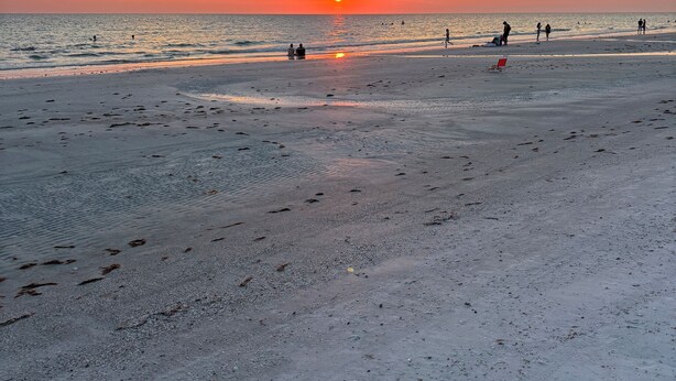En strand med fodspor, der fører ned til vandet, en solnedgang i baggrunden og et par personer, der går langs kysten.