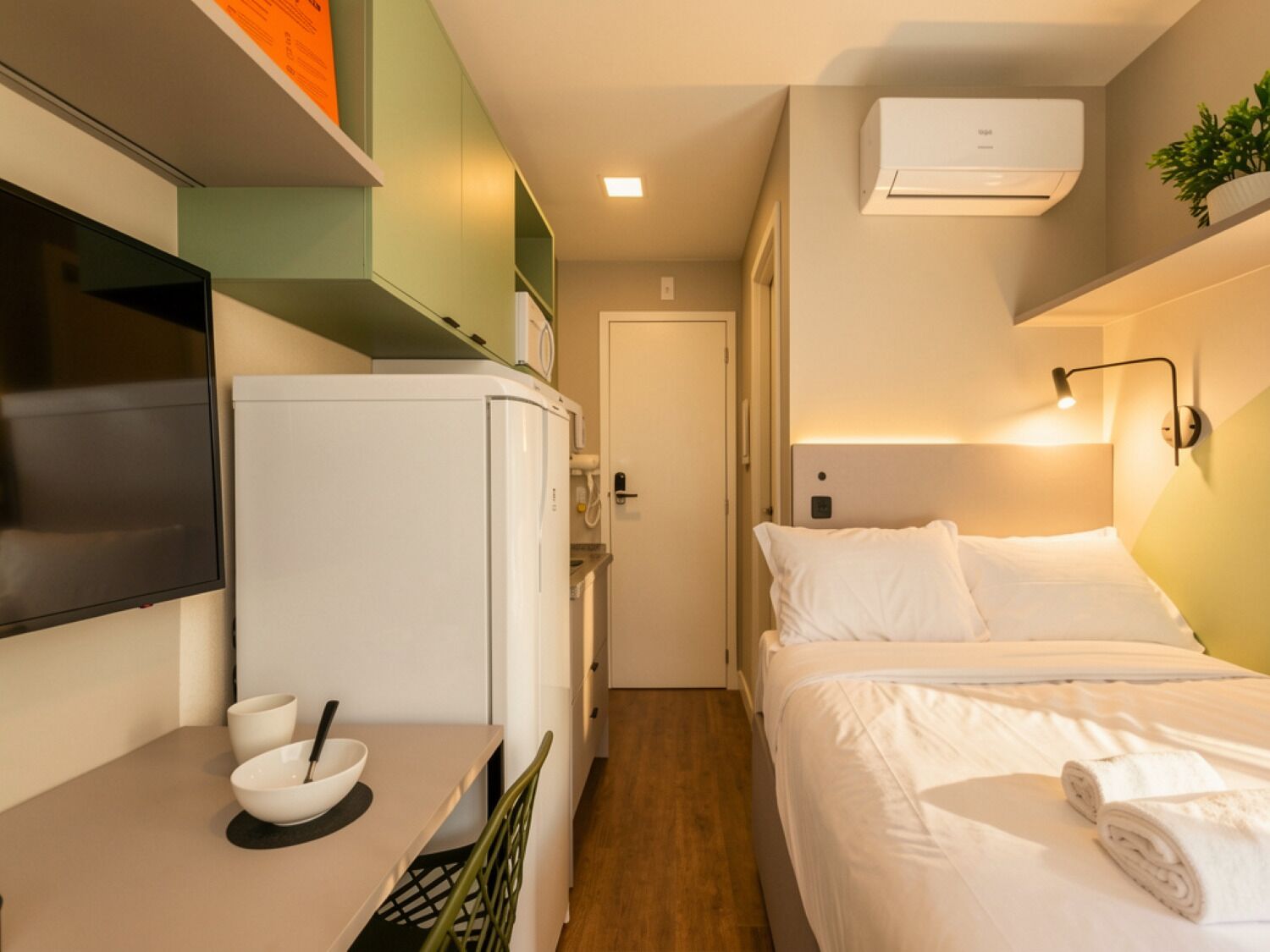 A compact hotel room with a bed, a small table with a bowl and spoon, a refrigerator, a TV mounted on the wall, and an air conditioning unit.