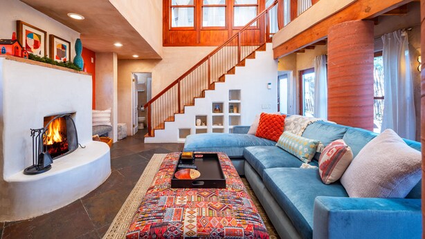 A spacious living room with a fireplace, a blue sofa, a patterned ottoman, and a wooden staircase. - Meticulously Designed Home with Lavish Furnishings (Santa Fe)