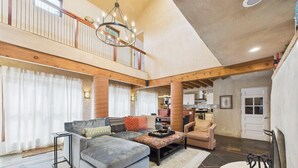 A spacious living area with a large sectional sofa, a coffee table, and a dining table. There is a kitchen in the background, a balcony with a railing, and a large window with curtains. - Meticulously Designed Home with Lavish Furnishings (Santa Fe)