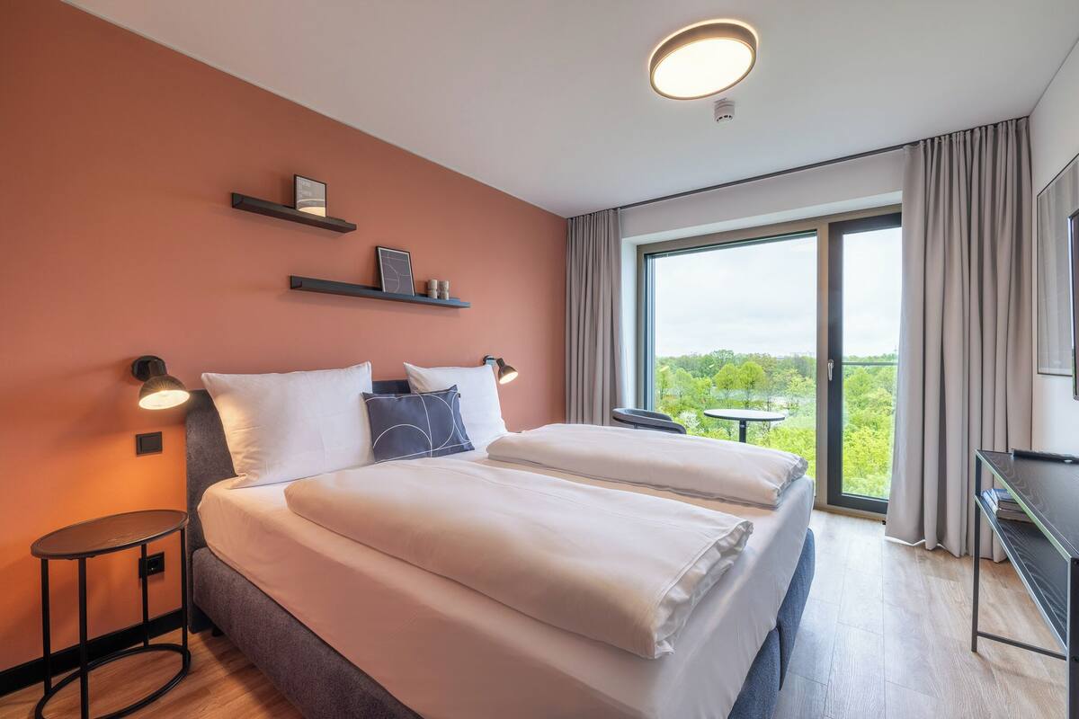 A double bed with white linens, two bedside tables, a wall-mounted shelf with items, a large window with curtains, and a ceiling light fixture.