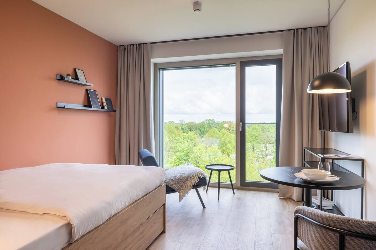 A modern hotel room with a large bed, a small table with a bowl and glass, a chair, a TV, and a balcony with a view of greenery.