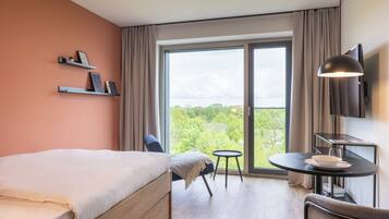 Habitación de hotel moderna con una cama grande, una mesita con un cuenco y una copa, una silla, un televisor y un balcón con vistas a zonas verdes.