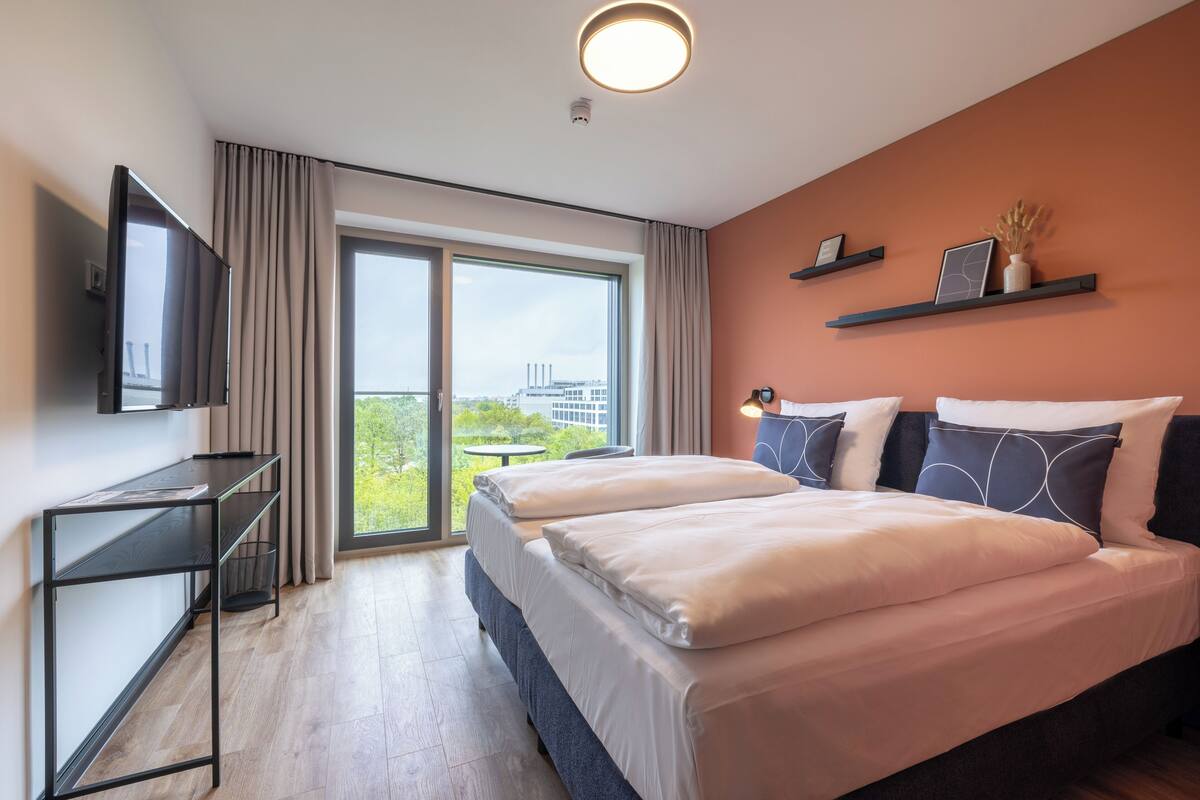 A modern hotel room with a large bed, a TV on a stand, a desk, and a balcony with a city view.