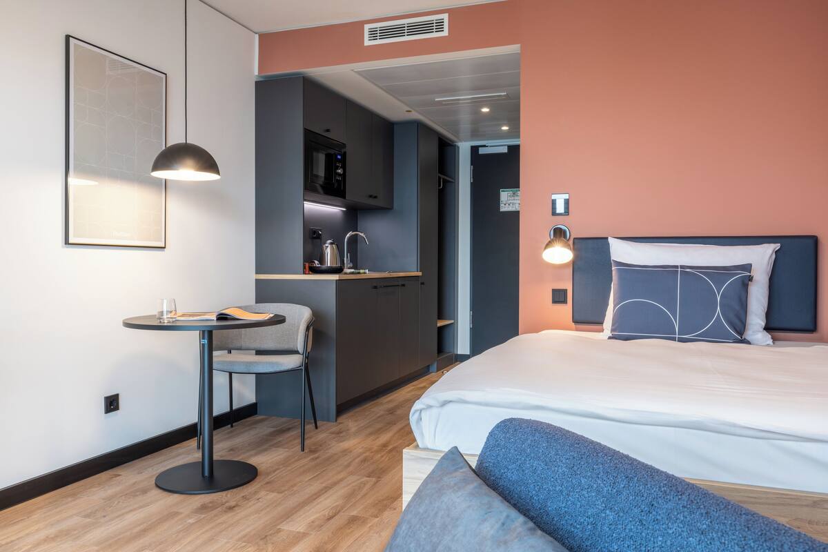 A modern hotel room with a bed, a desk, a chair, and a kitchenette.