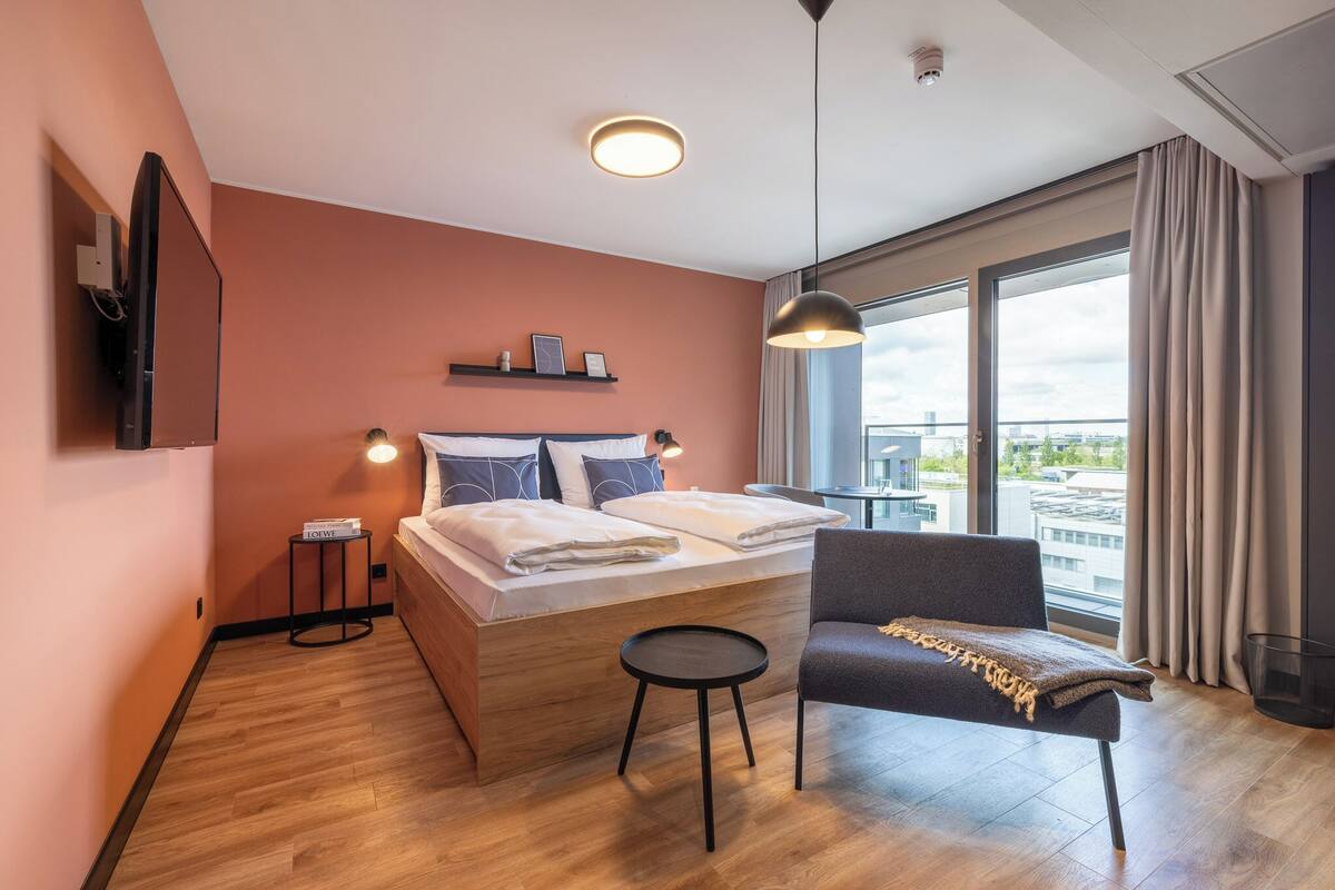 A modern hotel room with a large bed, a small table, a chair, a TV mounted on the wall, and a large window offering a city view.