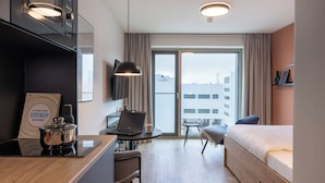 Ein modernes Hotelzimmer mit einer Küchenzeile, einem Laptop auf einem kleinen Tisch, einem Bett, einem Stuhl und Blick auf die Stadt durch das Fenster.