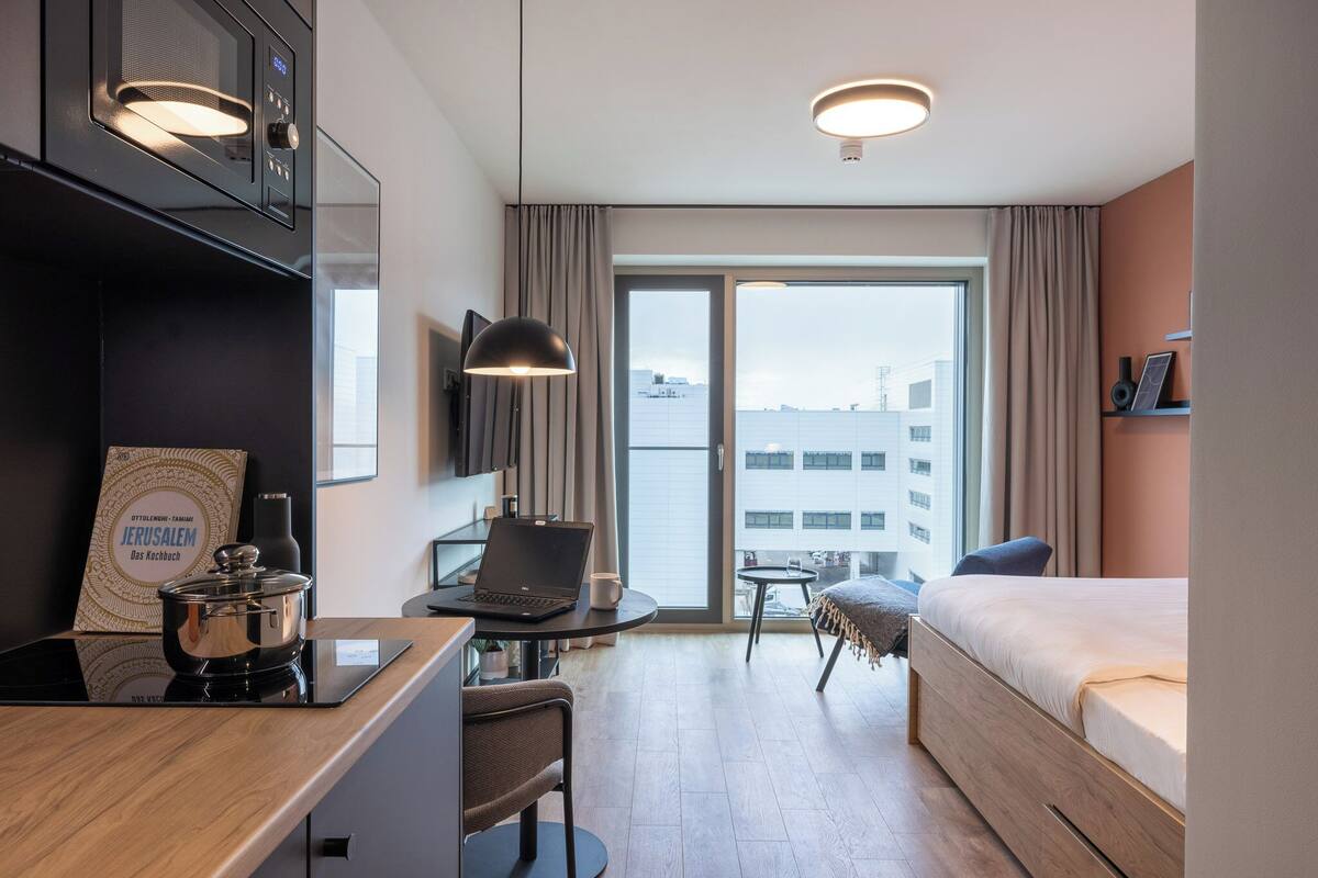 A modern hotel room with a kitchenette, a laptop on a small table, a bed, a chair, and a view of the city through the window.
