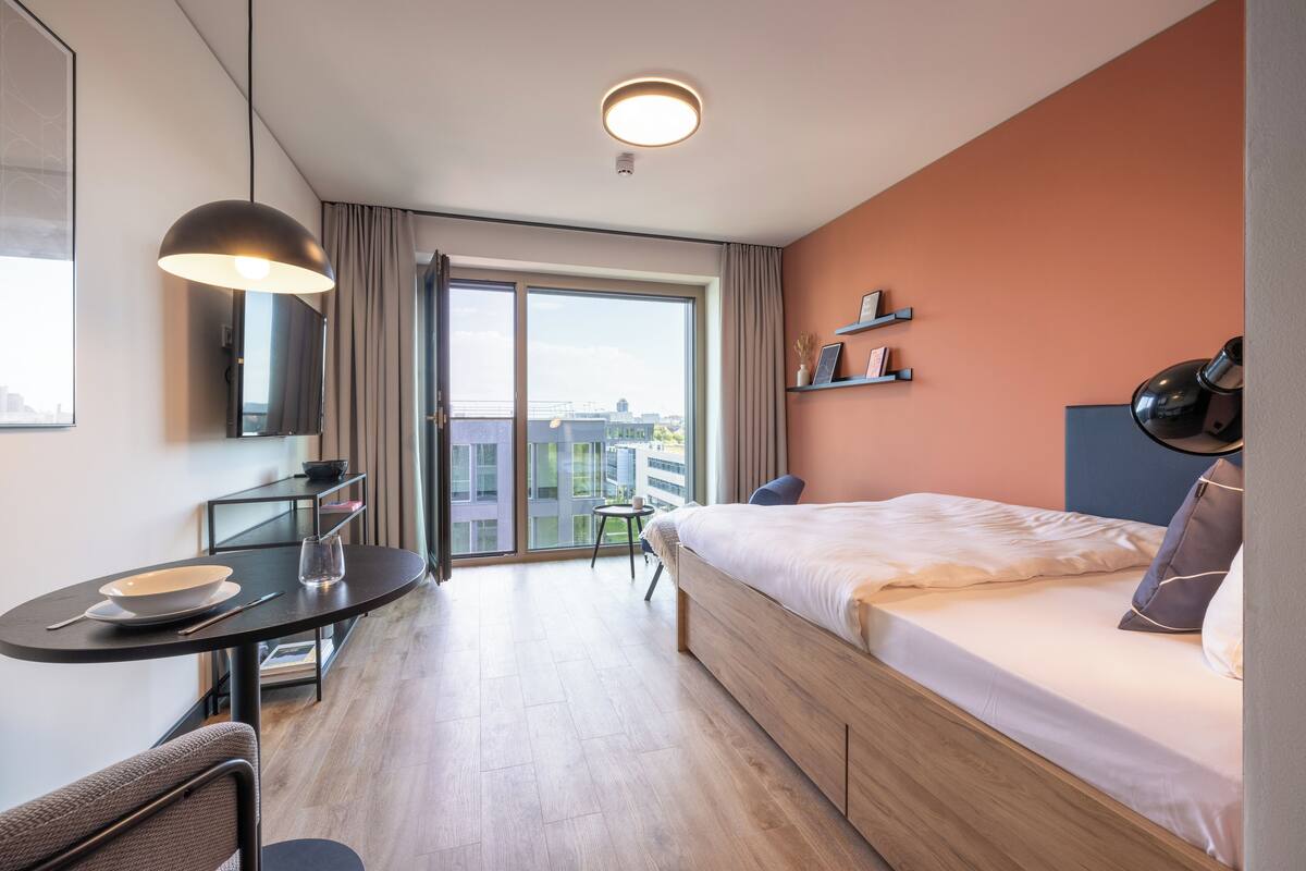 A modern hotel room with a bed, a desk with a chair, a small table, and a large window offering a city view.