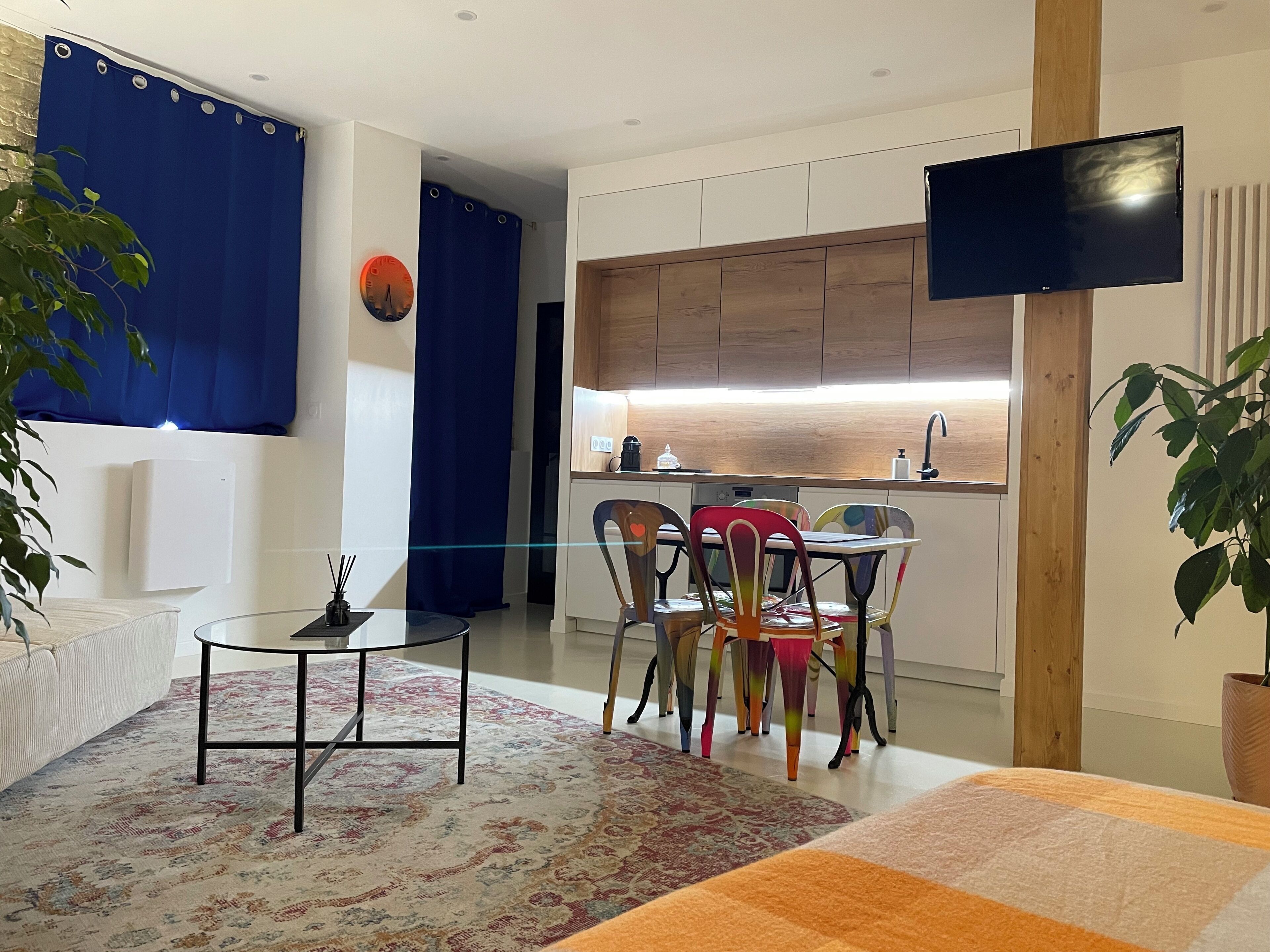 A modern living room with a glass coffee table, colorful chairs, a sofa, a flat-screen TV, and a kitchen area with wooden cabinets and a built-in oven.