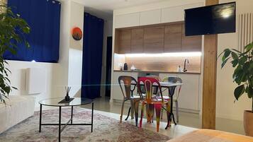 A modern living room with a glass coffee table, colorful chairs, a sofa, a flat-screen TV, and a kitchen area with wooden cabinets and a built-in oven.