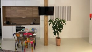 A modern kitchen with wooden cabinets, a built-in oven, a dining table with colorful chairs, a potted plant, and a mounted TV. - Studio 11 bis – Facing Vichy Parks (Vichy)