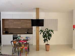 Una cocina moderna con armarios de madera, horno integrado, una mesa de comedor con sillas coloridas, una planta en maceta y un televisor montado.