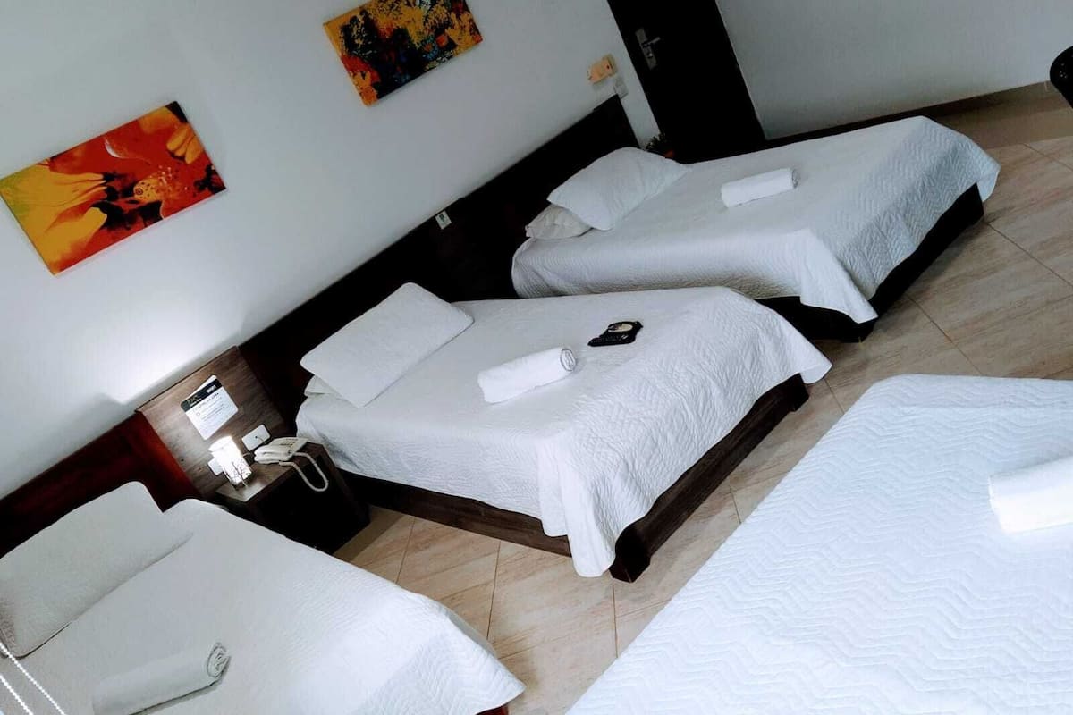 Quarto de hotel com três camas individuais, uma mesa de cabeceira com telefone e um quadro na parede.