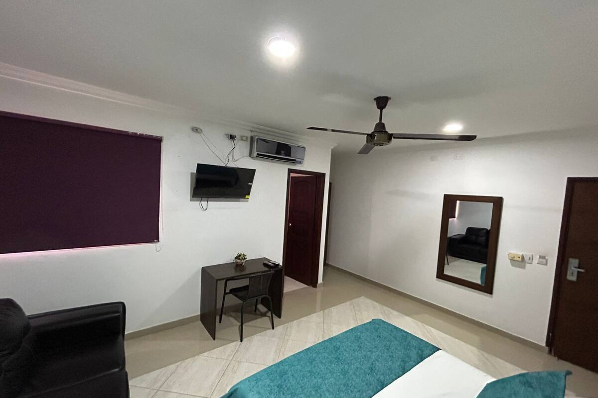 Quarto com cama, escrivaninha, cadeira, televisão, ventoinha no teto e janela com persianas.