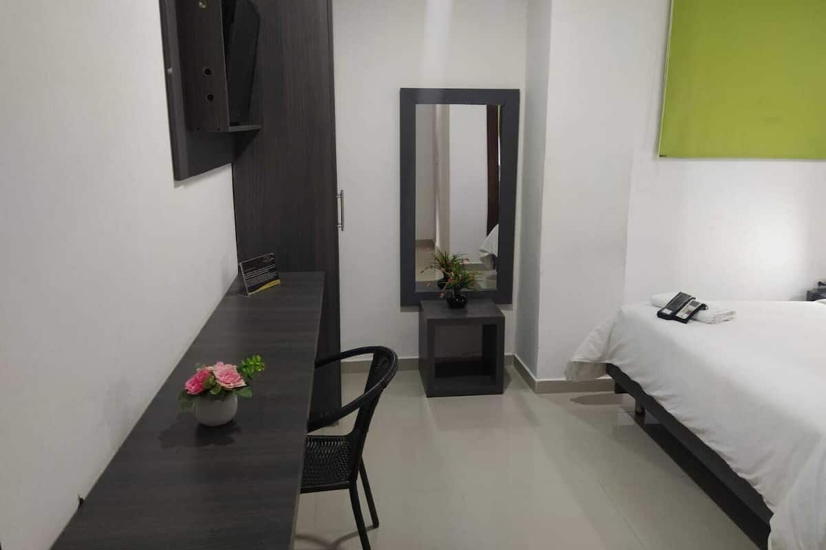 Quarto de hotel com cama, escrivaninha, cadeira, espelho e uma pequena mesa com um vaso de flores.