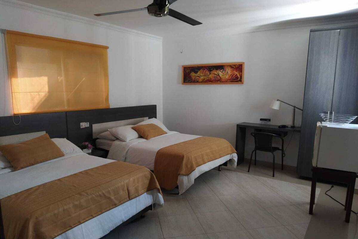 Quarto de hotel com duas camas, uma escrivaninha, uma cadeira, ventoinha no teto, um quadro e uma janela com persianas.