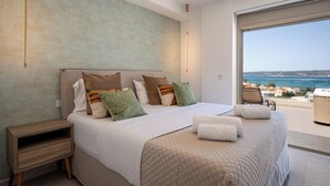 Une chambre moderne avec un grand lit, une table de chevet et une vue sur la mer, visible à travers une grande fenêtre.