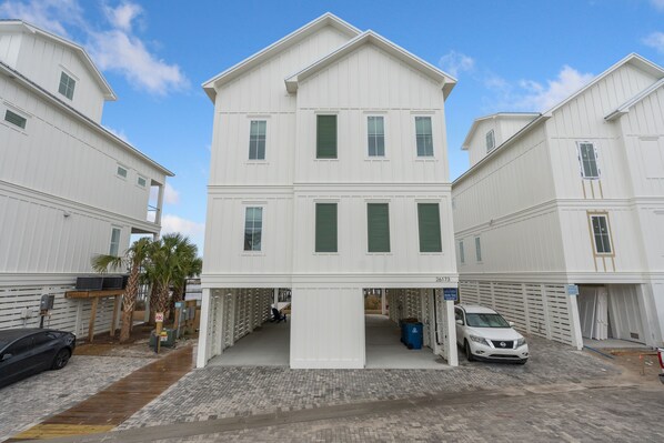 A white multi-story building with a central entrance, a covered parking area, and a row of lockers on the ground floor. - BRAND NEW HOME-Marlin Time-The Osprey-Signature Properties (Orange Beach)