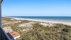 A beach view with a sandy shore, a clear sky, and some vegetation in the foreground.