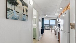 A narrow hallway leading to a balcony with a view, a kitchen area with white appliances, and a painting of palm trees on the wall.