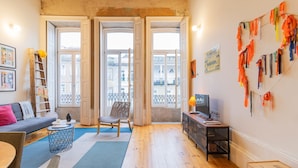 Ein gemütliches Wohnzimmer mit Sofa, Sessel, Couchtisch und TV-Stand. Zwei Fenster mit Vorhängen, eine Leiter und eine Wand mit Kalender und Lampe sind ebenfalls zu sehen.