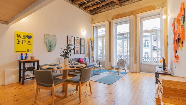 Ein gemütliches Zimmer mit einem Holzesstisch, Stühlen, einer Couch und einem großen Fenster mit Blick auf die Stadt.