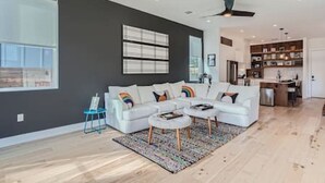 A modern living room with a white sectional sofa, a coffee table, and a rug. There is a kitchen area with wooden cabinets and a dining table. - Dip into the 512 Experience! 6 BDRMS W/Pool Mins to DT (Austin)