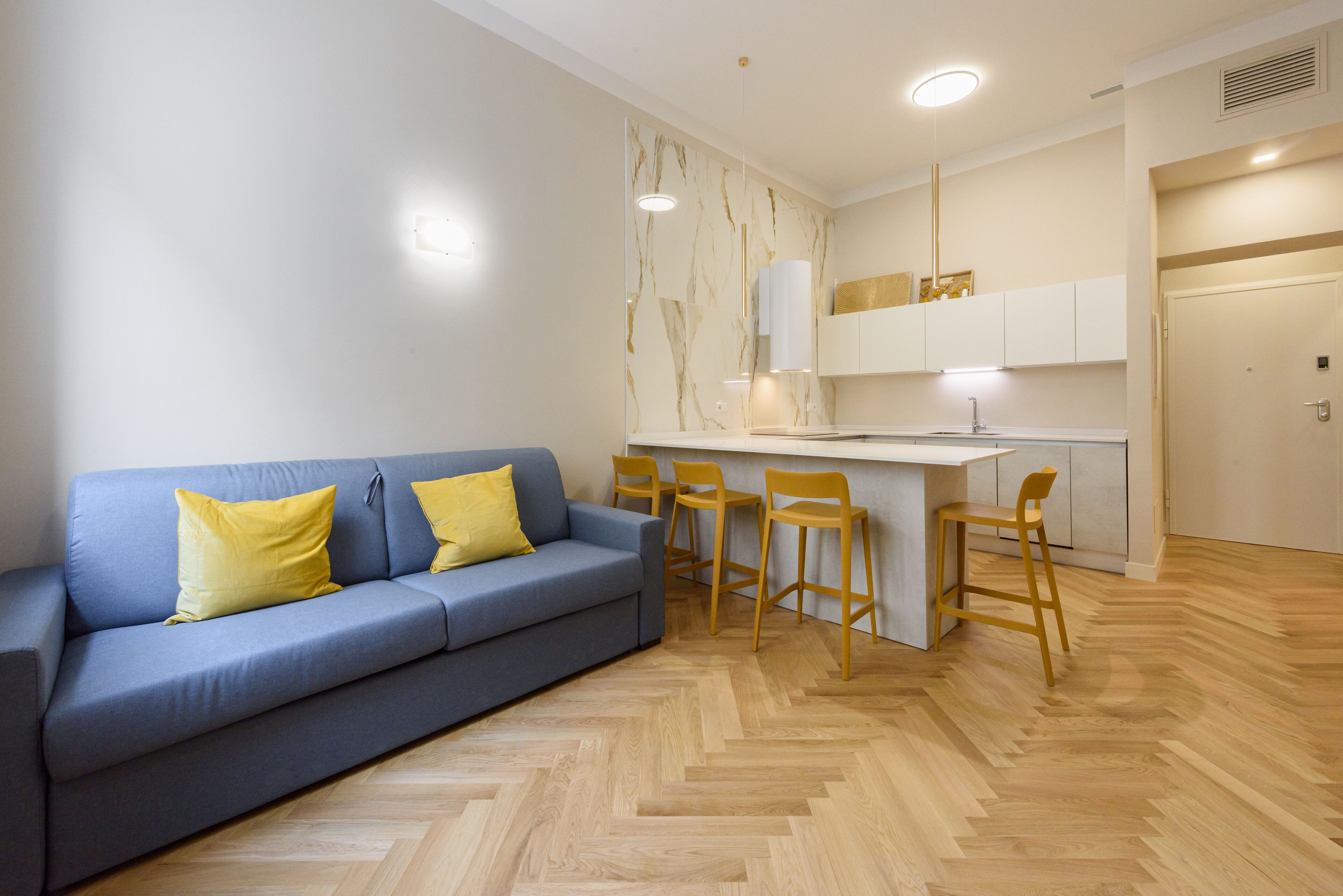 A modern living room with a blue sofa, yellow cushions, a wooden herringbone floor, and a kitchen area with a white countertop and wooden chairs.