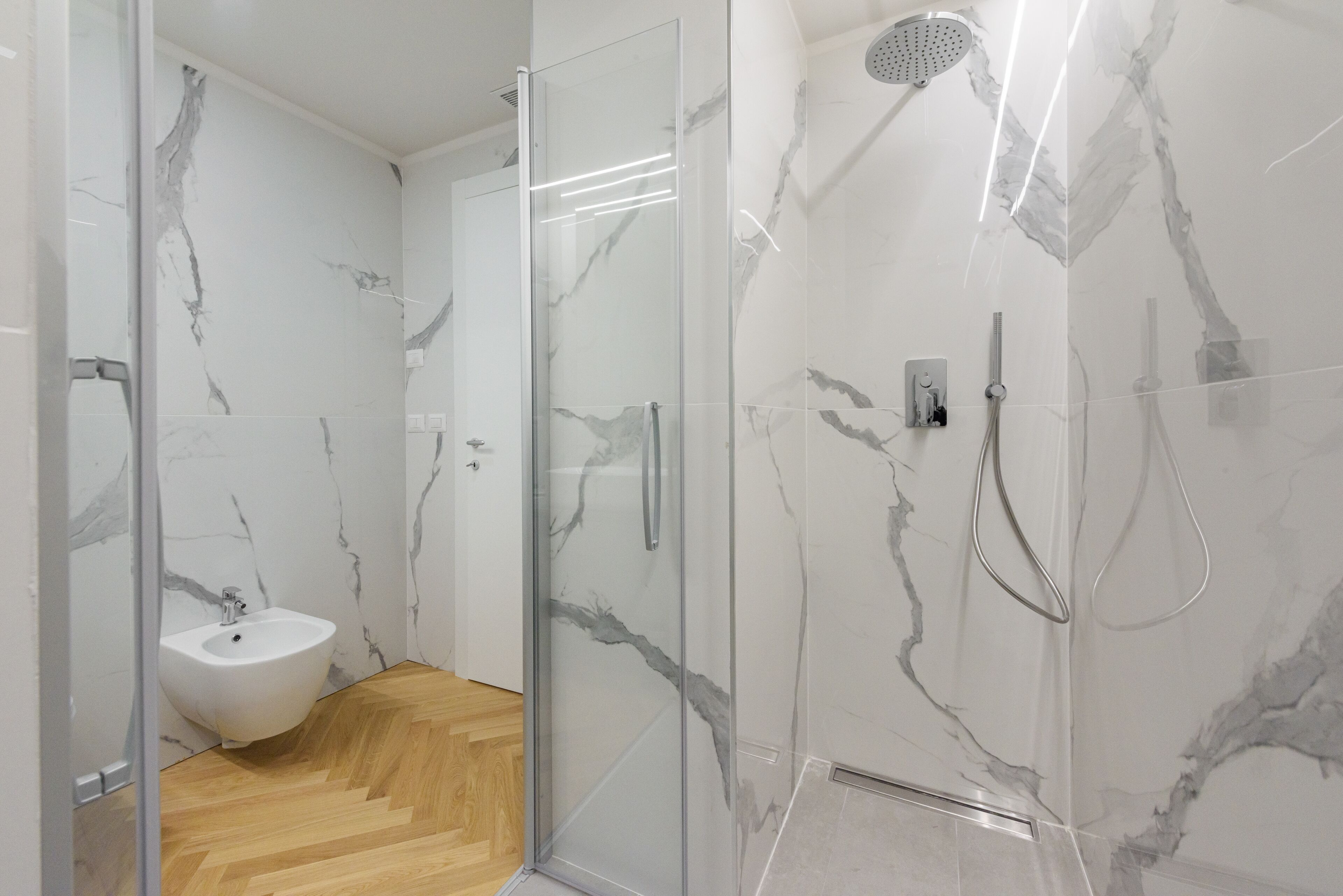A modern bathroom with a glass-enclosed shower, a white pedestal sink, and marble-patterned walls.