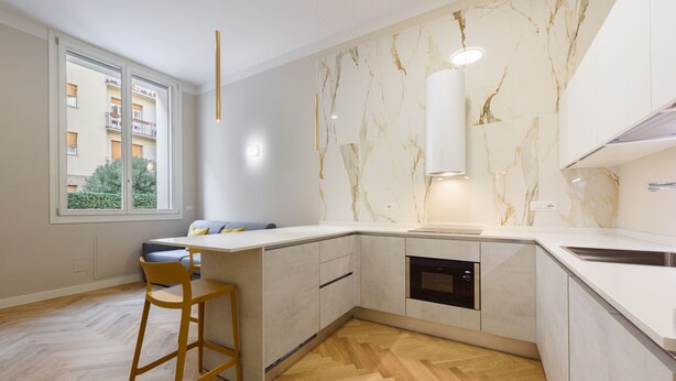 Une cuisine moderne avec un dosseret en marbre, un four encastré et une grande fenêtre donnant sur un immeuble d’habitation.