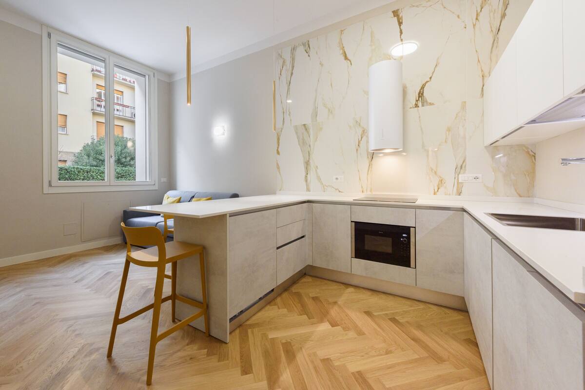 A modern kitchen with a marble backsplash, a built-in oven, and a large window offering a view of a residential building.