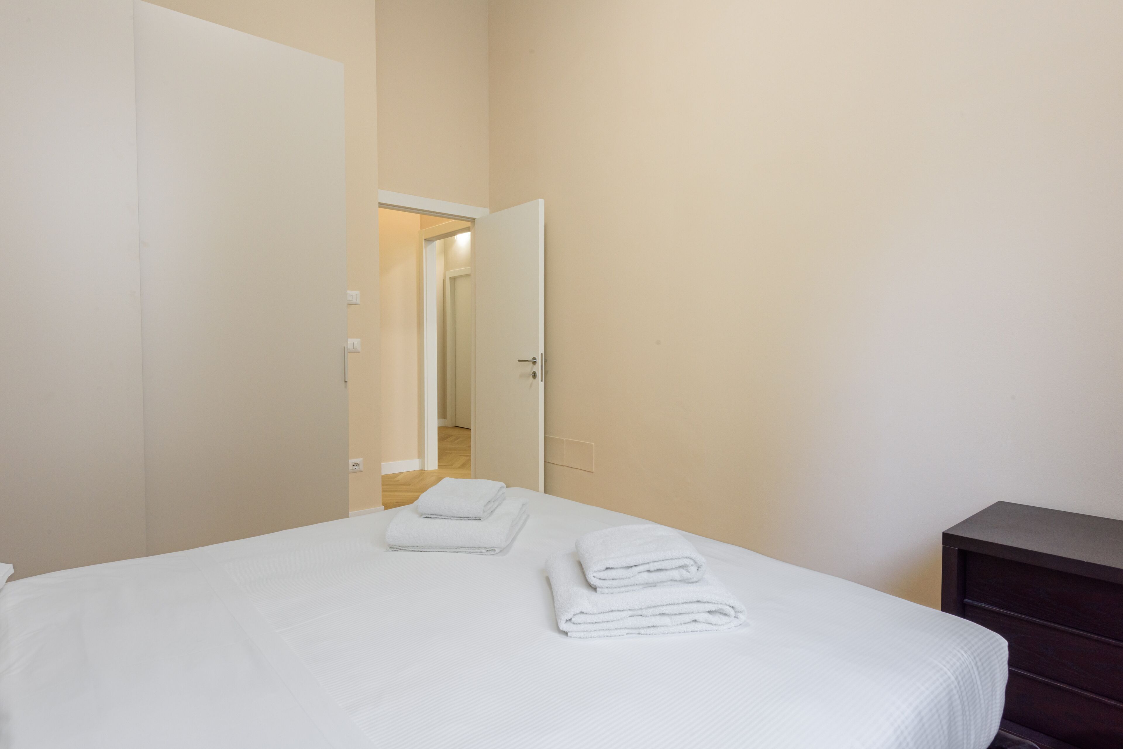 A hotel room with a bed, two neatly folded white towels, a dark wooden dresser, and a door leading to another room.
