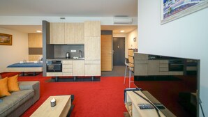 Een moderne woonkamer met een bank, een salontafel en een televisie. Er is een keukenhoek met kastjes en een fornuis.