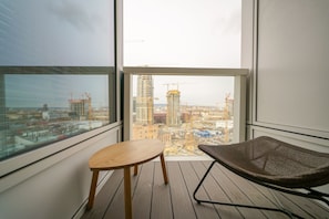 Un balcon avec une table en bois et une chaise en osier, donnant sur un paysage urbain où l’on aperçoit des grues de construction.