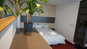 Een kleine hotelkamer met een bed, een plant in pot, een nachtkastje en een ingebouwde plank met toiletartikelen.