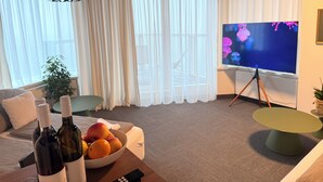 Een woonkamer met een bank, een salontafel met fruit en wijnflessen, een televisie op een statief en dunne gordijnen.