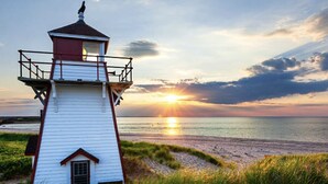 A white lighthouse with a red roof and balcony, situated on a grassy beach with the sun setting in the background.