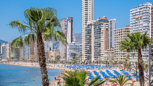 A beachside cityscape with high-rise buildings, palm trees, and a sandy beach with blue umbrellas.