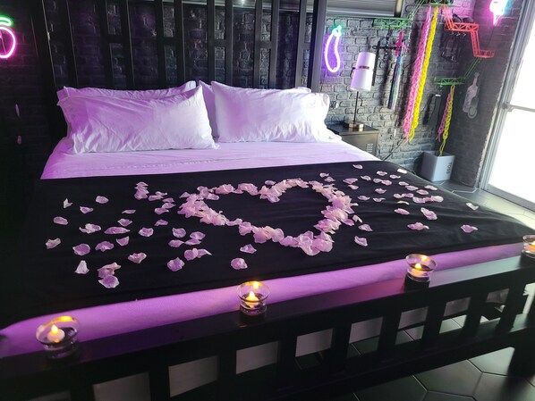 A bed with a heart-shaped arrangement of rose petals and candles, a brick wall, and neon signs.
