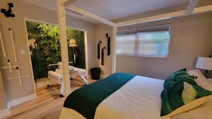 A bedroom with a bed, a window with blinds, a bedside lamp, and a view of a green outdoor area.