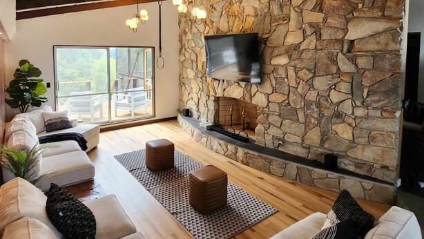 A modern living room with a stone wall, a fireplace, and a large TV mounted on the wall. There is a sofa set with cushions and a coffee table.