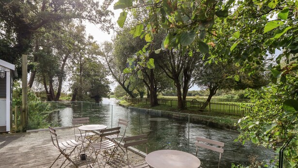 Terraza a la orilla del río con sillas y mesas, rodeada de árboles y una valla.