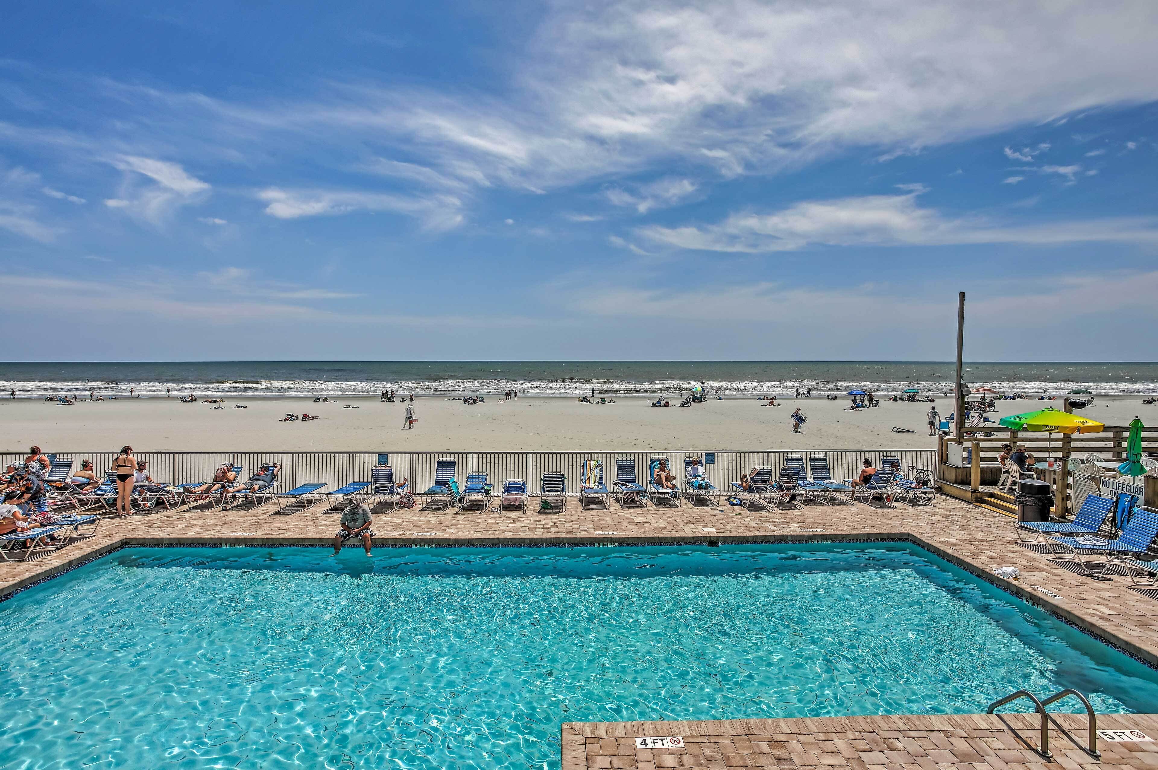 A pool area with clear blue water, a beach in the background, and a wooden deck with lounge chairs.