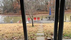 Ein überdachter Außenbereich mit Betonboden, einer Holzterrasse und Blick auf einen Teich mit Stühlen und einem Tisch.