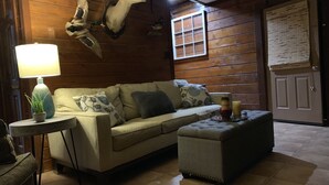 Ein gemütliches Zimmer mit Holzwand, einer Couch, einem Hocker, einer Lampe und einem aufgehängten Hirschkopf.