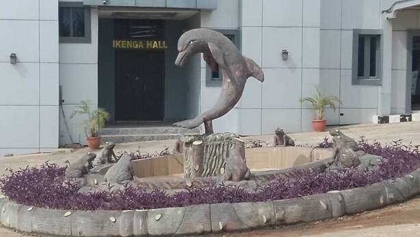 A dolphin sculpture in a circular planter with purple flowers, in front of a building labeled "IKENGA HALL".