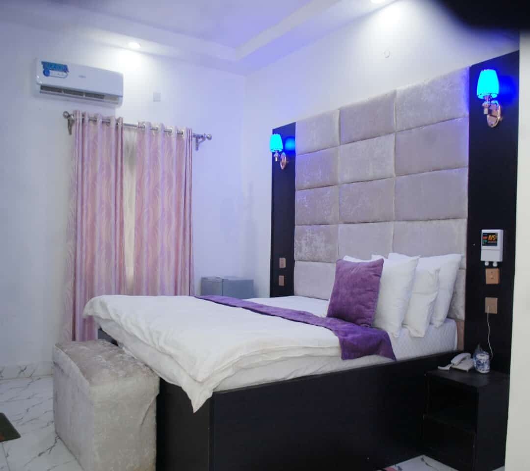 A bedroom with a bed, a nightstand, an air conditioning unit, a wall-mounted lamp, and a curtain.