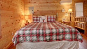 Un dormitorio de cabaña de madera con ambiente acogedor, colcha a cuadros, cabecera con motivos de ciervo y una pequeña mesa con lámpara.